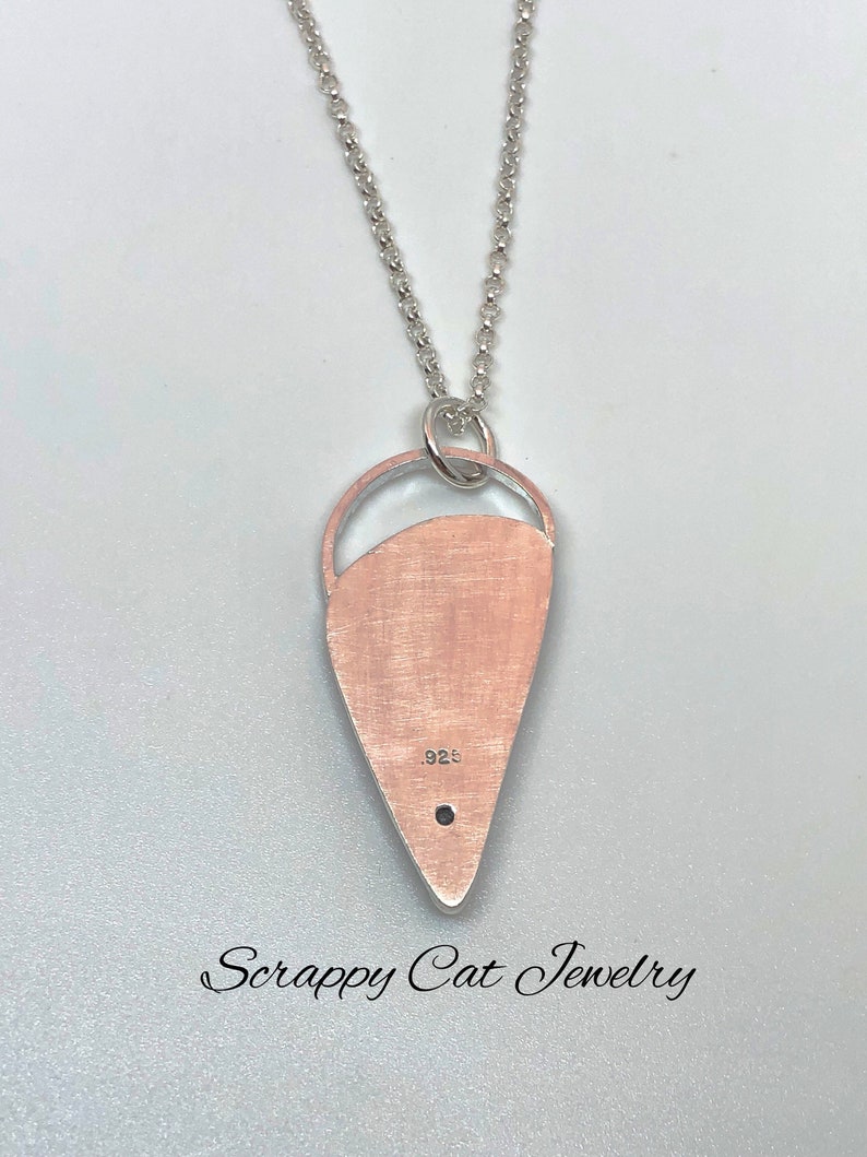 May include: A silver necklace with a teardrop-shaped pendant. The pendant has a cutout design and is marked with "925". The necklace is from "Scrappy Cat Jewelry".