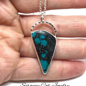 May include: A silver necklace with a turquoise and black stone pendant. The pendant is shaped like a teardrop and has a silver bail. The necklace is on a silver chain.