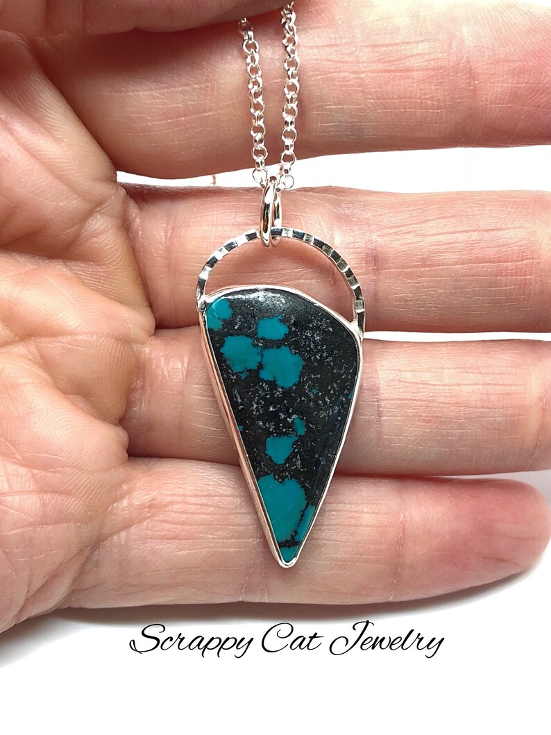 May include: A silver necklace with a turquoise and black stone pendant. The pendant is shaped like a teardrop and has a silver bail. The necklace is on a silver chain.