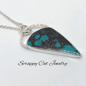 May include: A turquoise and black stone pendant with a silver setting and a silver chain. The pendant is shaped like a teardrop and has a circular design around the stone. The text "Scrappy Cat Jewelry" is visible below the pendant.