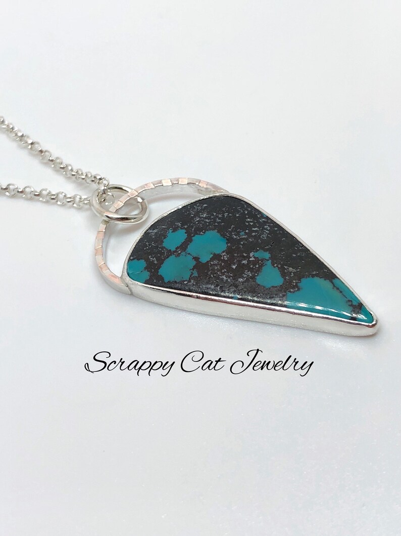 May include: A turquoise and black stone pendant with a silver setting and a silver chain. The pendant is shaped like a teardrop and has a circular design around the stone. The text "Scrappy Cat Jewelry" is visible below the pendant.