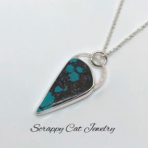 May include: A silver chain necklace with a turquoise and black stone pendant. The pendant is set in a silver bezel and has a silver ring around it. The necklace is on a white background. "Scrappy Cat Jewelry" is written in cursive at the bottom of the image.