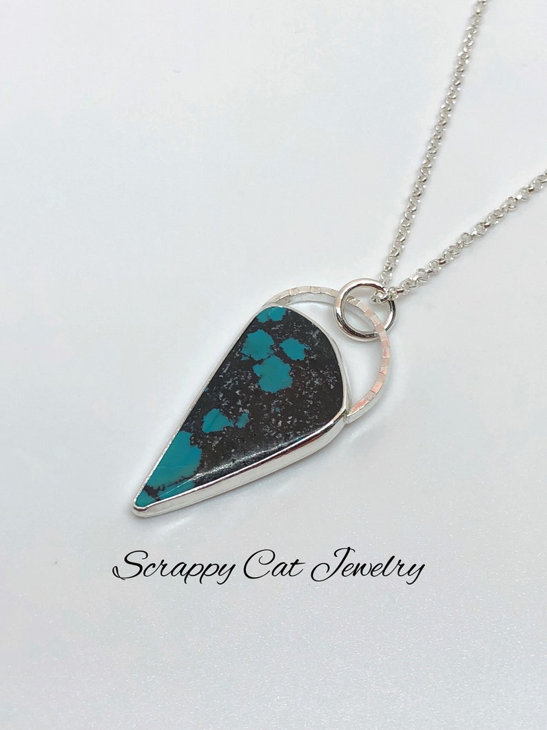 May include: A silver chain necklace with a turquoise and black stone pendant. The pendant is set in a silver bezel and has a silver ring around it. The necklace is on a white background. "Scrappy Cat Jewelry" is written in cursive at the bottom of the image.