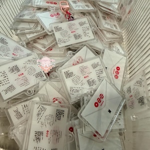 May include: A collection of clear acrylic keychain charms shaped like envelopes. Each charm features a white envelope design with a red circular logo, a small black logo, and printed text resembling a handwritten letter. Some charms include a pink clasp.