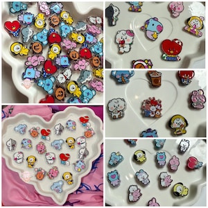 May include: A collection of colorful cartoon character charms in various shapes and designs. The charms are primarily in shades of blue, pink, yellow, and red, with black outlines. They are arranged on white, heart-shaped trays.