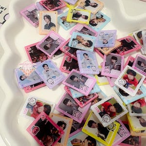 May include: A collection of colorful, small, rectangular charms featuring images of people. The charms have various colored borders, including pink, yellow, and blue. Some charms have text, including the names "SUGA", "J-HOPE", and "JIN".