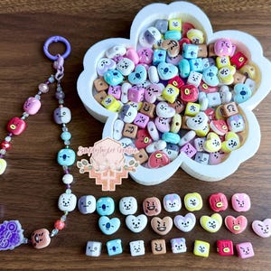 May include: A beaded lanyard with a purple ring and colorful character-themed beads, a white flower-shaped tray filled with assorted character beads, and a collection of loose square and heart-shaped character beads with the text Simply Taylor Creation.