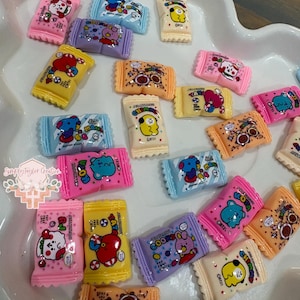 May include: A collection of small, rectangular, candy-shaped resin charms featuring various colorful BT21 character graphics. Each charm displays unique illustrations of characters like Tata, Chimmy, Koya, and Cooky in vibrant hues of pink, blue, yellow, and orange.