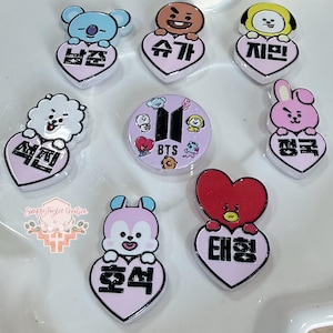 May include: A collection of eight small, glossy, heart-shaped charms featuring cartoon characters and Korean text. Seven charms depict characters holding hearts with names, while one central circular charm displays the BTS logo and small icons.