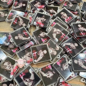 May include: A collection of square, clear acrylic keychains featuring printed portraits of various individuals. Each keychain displays a black and white photograph of a person with red circular graphic accents and text overlays.