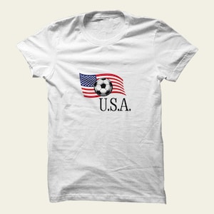 May include: White t-shirt with a graphic of a soccer ball incorporated into the American flag design. The text "U.S.A." is printed below. The shirt is made of a soft material and has a crew neck.