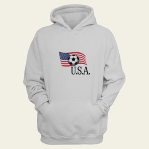 May include: White hoodie with a graphic of the American flag, a soccer ball, and the text "U.S.A.". The sweatshirt has a front pocket and a drawstring hood. A casual, sporty garment.
