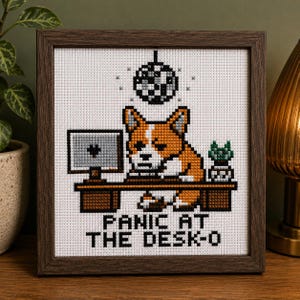 May include: Cross-stitch artwork in a dark brown frame. The image features a corgi at a desk with a computer, a disco ball, and the text "PANIC AT THE DESK-O". The corgi is orange and white, and the desk is brown.