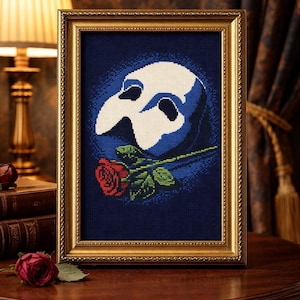 May include: Framed cross-stitch art featuring the Phantom of the Opera mask in white and blue, with a red rose. The artwork is set against a dark blue background and framed in gold. The piece is displayed on a wooden table.