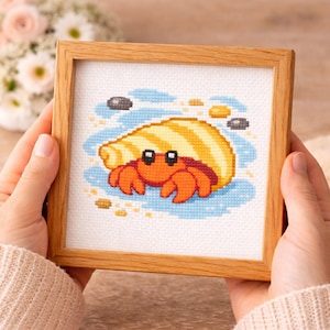 May include: A framed cross-stitch art piece featuring a cartoon hermit crab. The crab is orange with a yellow and white striped shell, set against a blue and sand-colored background. The frame is made of light brown wood.