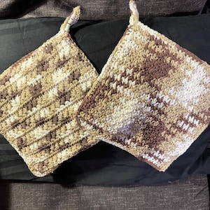 May include: Two handmade crochet potholders in shades of brown, beige, and white. Each square potholder has a loop for hanging. The textured stitch creates a visually appealing pattern. These kitchen accessories are perfect for protecting hands from heat.