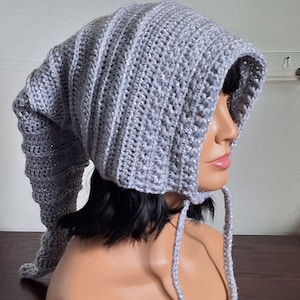 May include: A gray, handmade crochet hat with a long, pointed tip and a hood. The hat has a sparkly, textured appearance and ties under the chin with a matching cord. The hat is a unique, whimsical accessory.