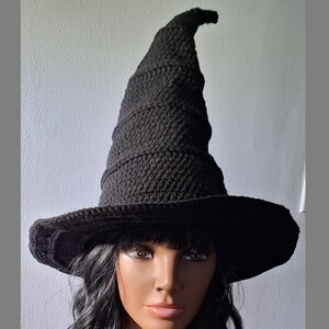 May include: A black crocheted witch hat with a tall, pointed crown and a wide brim. The hat is worn by a person with dark hair.