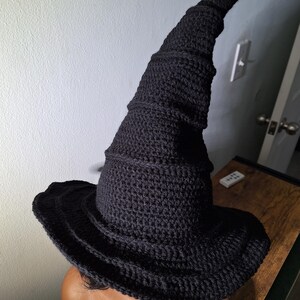 May include: A black crocheted witch hat with a tall, pointed crown and a wide brim. The hat is made with a textured stitch and has a spiral design.