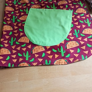 May include: A burgundy apron with a green pocket. The apron is patterned with tacos, cacti, lime slices, and tortilla chips. The pocket is a solid green color. The apron is designed for kitchen use.