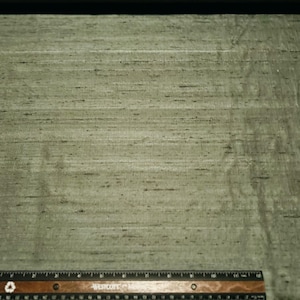 May include: Close-up of a textured, olive green fabric with subtle horizontal lines and dark flecks. A wooden ruler with inch markings is at the bottom. The fabric has a slight sheen.