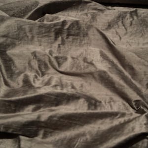 Op de afbeelding: Close-up van een gedrapeerde stof met een glanzende, metallic uitstraling. De stof is een gedempte, donkergrijze kleur met zichtbare plooien en texturen, waardoor een gevoel van diepte en beweging ontstaat. Het materiaal lijkt zacht en vloeiend.