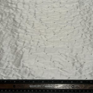 May include: White fabric with a textured surface and scattered, small, shiny embellishments. A black ruler with white markings is at the bottom of the image. The fabric appears to be a decorative textile.