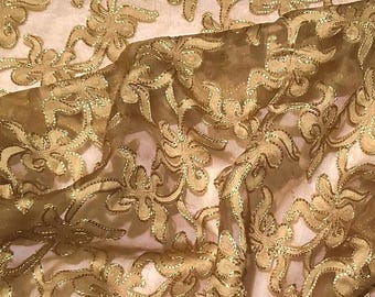 May include: Close-up of a sheer fabric with a floral pattern. The fabric is a golden colour with gold metallic thread outlining the floral design. The background is a light beige colour.