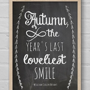 Puede incluir: Un letrero de pizarra enmarcado con la cita "Autumn, the year's last loveliest smile" en escritura blanca. El letrero tiene un marco de madera marrón claro y está suspendido por una cuerda natural. El fondo es una pared de ladrillos blancos.