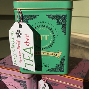 May include: A stack of teal and pink tea tins with gold trim. The teal tin has a tag that reads "Merry Christmas to a wonderful TEAcher." The tins are labeled "HT Blends Fine Teas" and "Peppermint Herbal."