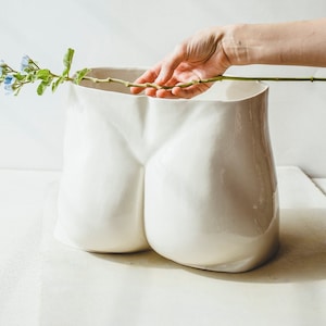 May include: A white ceramic vase with a unique form, resembling a pair of buttocks. A hand is placing a stem of blue flowers into the vase. The vase is on a white surface, with a white background.