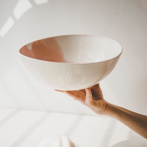 May include: A white ceramic bowl with a pink rim is held in a hand against a white background. A white pumpkin sits on the surface below the bowl.