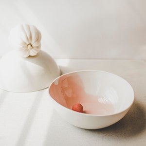 May include: A white ceramic bowl with a pink glaze on the inside. A single red lychee sits in the bowl.