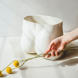 Może przedstawiać: Biała ceramiczna doniczka w kształcie ludzkiego tyłka, z ręką trzymającą gałązkę suszonych żółtych kwiatów.