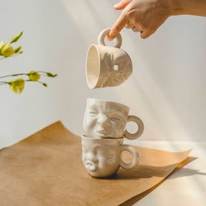 Może przedstawiać: Trzy białe ceramiczne kubki z twarzami. Kubki są ułożone jeden na drugim, a górny kubek jest trzymany przez rękę. Kubki znajdują się na brązowej papierowej powierzchni.