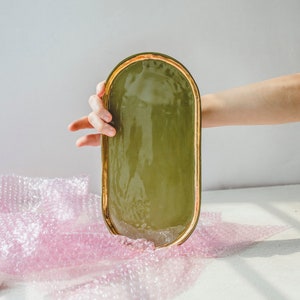 以下が含まれることがあります： 金色の縁取りのある緑色の楕円形のセラミックトレイ。トレイは手で持たれています。