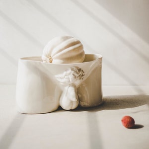 Puede incluir: Un macetero de cerámica blanca con forma de cuerpo humano con una calabaza blanca en su interior. Un pequeño fruto de lichi rojo se encuentra en la mesa en primer plano.