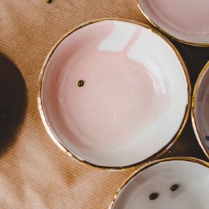 Może przedstawiać: Cztery małe ceramiczne miski ze złotymi obw&oacute;dkami. Miski są w r&oacute;żnych kolorach, w tym r&oacute;żowym, białym, niebieskim i szarym. Każda miska ma inny wz&oacute;r.
