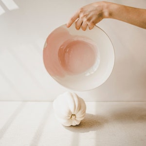 May include: A white ceramic bowl with a pink interior is being held upside down over a white gourd. The bowl and gourd are on a white surface.