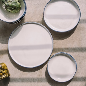 May include: Four white ceramic plates with blue trim, arranged on a light gray surface. The plates are of varying sizes, with the largest plate in the center.