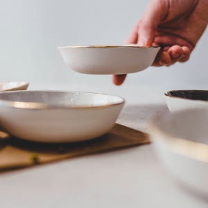 Może przedstawiać: Zestaw pięciu białych ceramicznych miseczek ze złotymi obw&oacute;dkami. Miseczki są ułożone w rzędzie na brązowej powierzchni. Jedna miseczka jest trzymana w ręku.