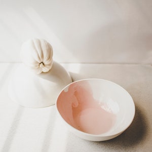 May include: A white ceramic bowl with a pink interior and a white ceramic lid with a white gourd on top.