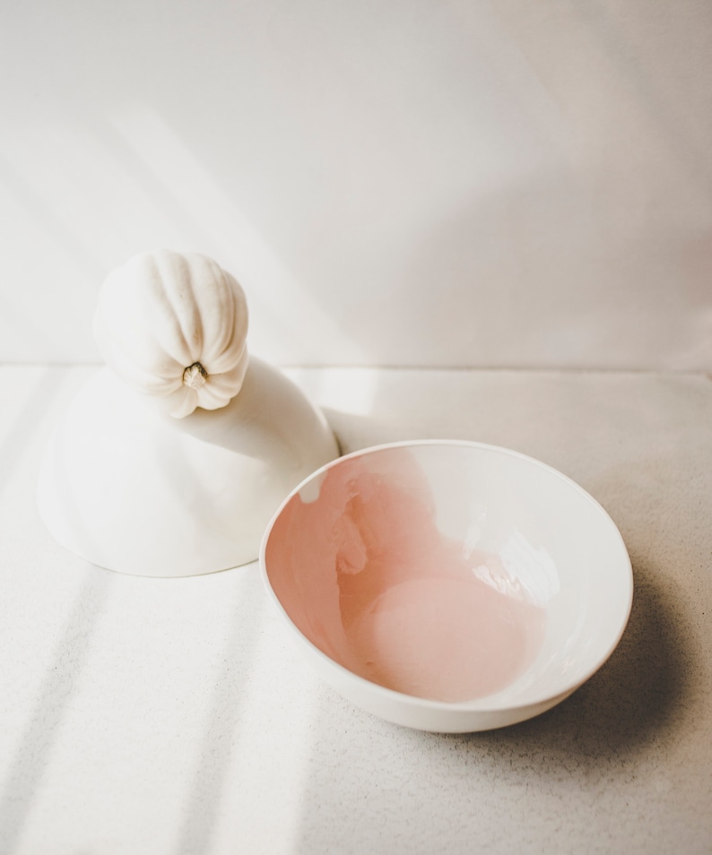 May include: A white ceramic bowl with a pink interior and a white ceramic lid with a white gourd on top.