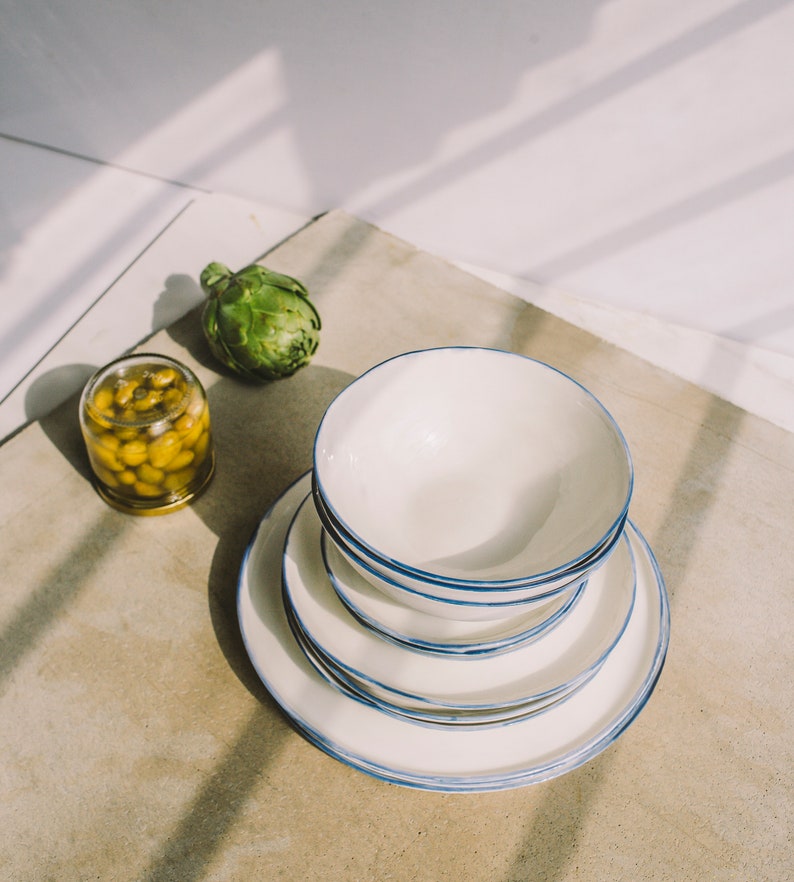 May include: A stack of four white ceramic plates with a blue rim. The plates are stacked on a light brown surface. A small jar of olives and a green artichoke are also on the surface.
