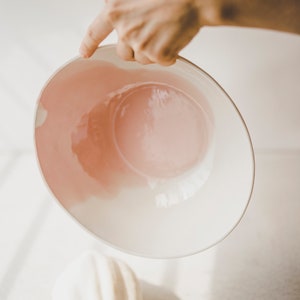 May include: A white ceramic bowl with a pink glaze inside. The bowl is being held by a hand.