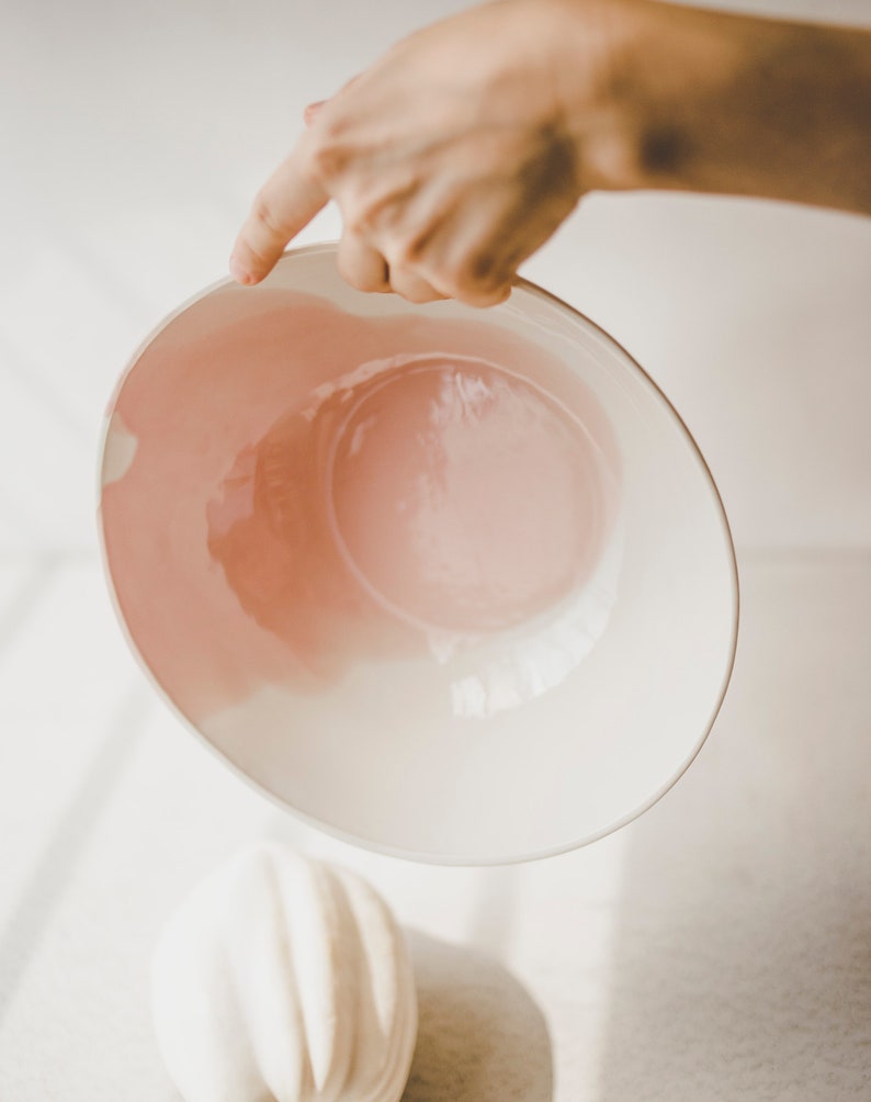 May include: A white ceramic bowl with a pink glaze inside. The bowl is being held by a hand.