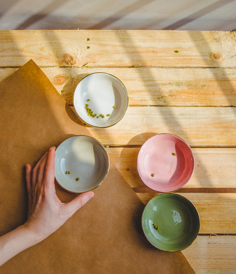May include: Four small ceramic bowls with gold rims, each containing a few green mung beans. The bowls are on a wooden surface with a brown paper sheet underneath.