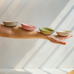 May include: Four ceramic bowls stacked on a hand, starting with a light gray bowl, then a pink bowl, a green bowl, and a white bowl.
