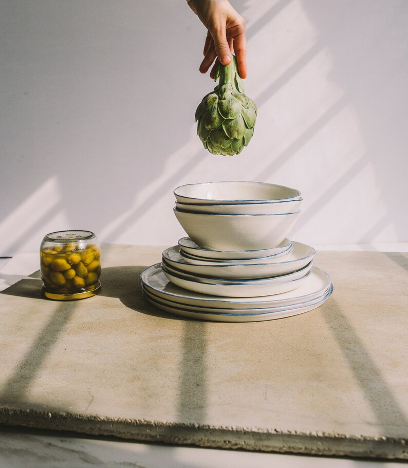 May include: A stack of white ceramic plates with blue trim, a jar of green olives, and a green artichoke held by a hand.