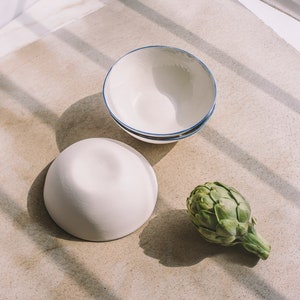 May include: Two white ceramic bowls, one with a blue rim, and a green artichoke on a light brown surface.